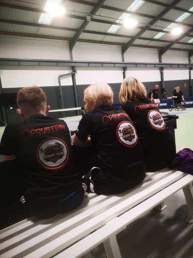 View of the back of 3 people sat down watching pickleball, wearing the Hybrid Team T-shirts